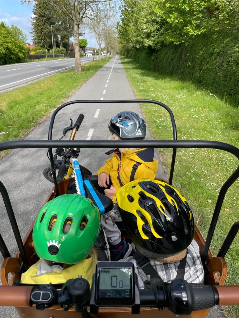 Tre børn sidder i ladkassen på en ladcykel fra EventyrCykler, mens der køres på en cykelsti